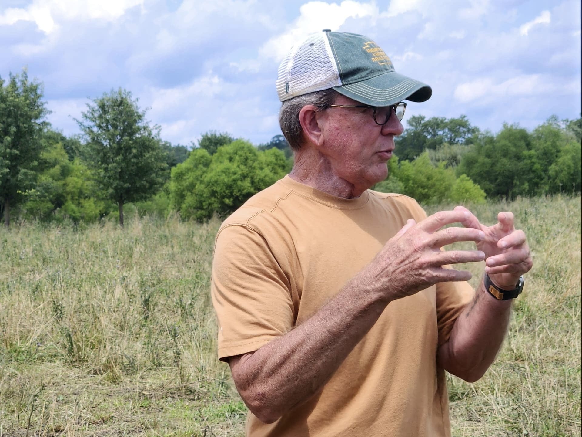A man in a tan shirt and green cap discusses sustainable farming practices while standing in a grassy field at Tyner Pond Farm
