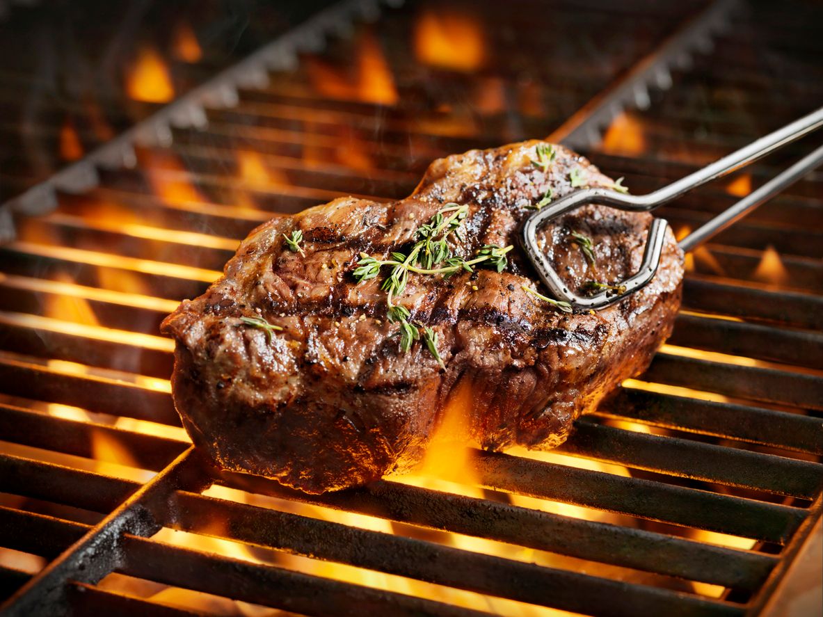 A grass-fed beef sirloin steak being cooked on a grill, shown with visible flames and seasoning.