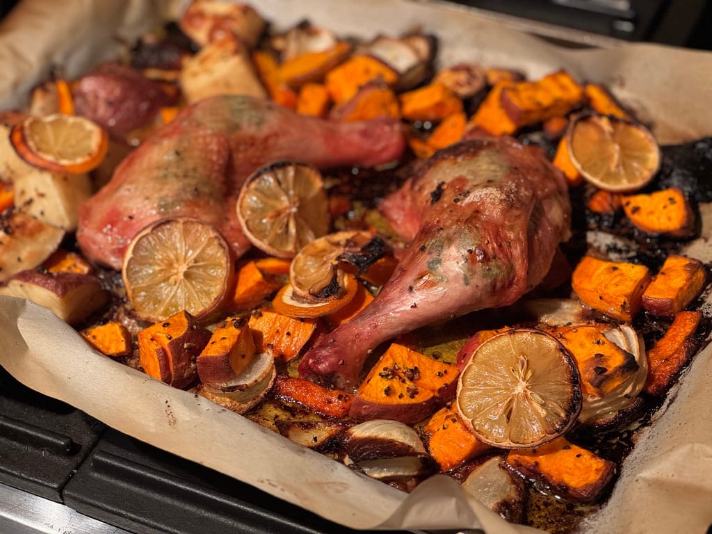 Roasted chicken quarters with crispy skin, surrounded by caramelized sweet potatoes, onions, carrots, and lemon slices on a parchment-lined sheet pan.
