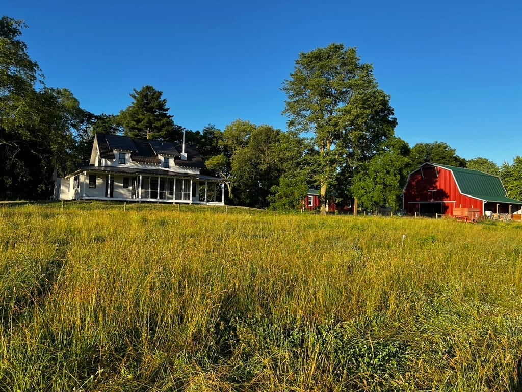 A farmhouse and red barn at Tyner Pond Farm surrounded by green pasture in the morning sunlight