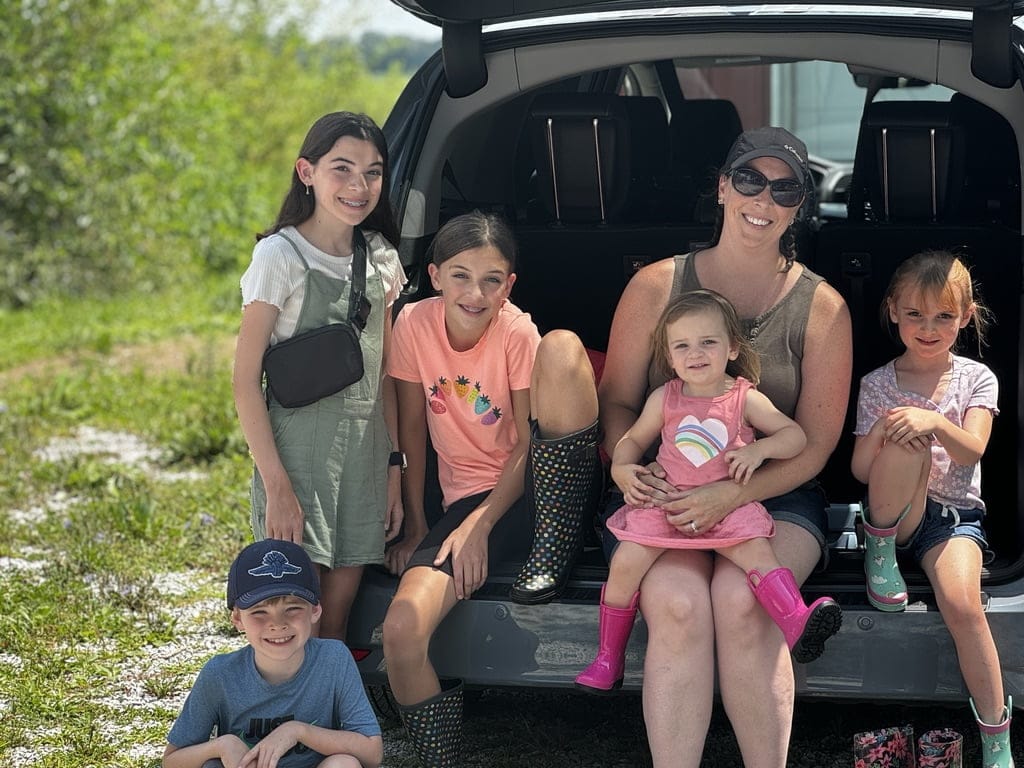 A homeschooling family sits in the back of their vehicle at Tyner Pond Farm, enjoying a sunny day on the farm. The group includes a mother and her five children, all smiling and dressed for outdoor exploration.