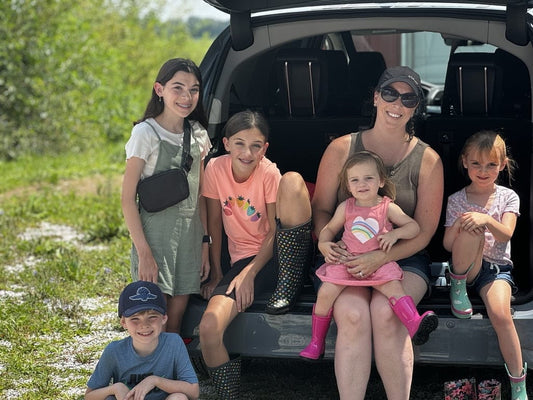 A homeschooling family sits in the back of their vehicle at Tyner Pond Farm, enjoying a sunny day on the farm. The group includes a mother and her five children, all smiling and dressed for outdoor exploration.