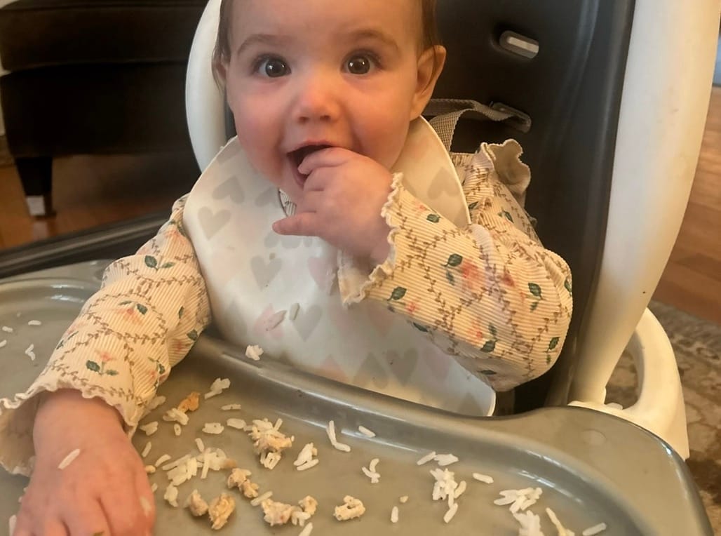A baby sitting in a high chair, eating rice and small pieces of ground chicken during baby-led weaning.