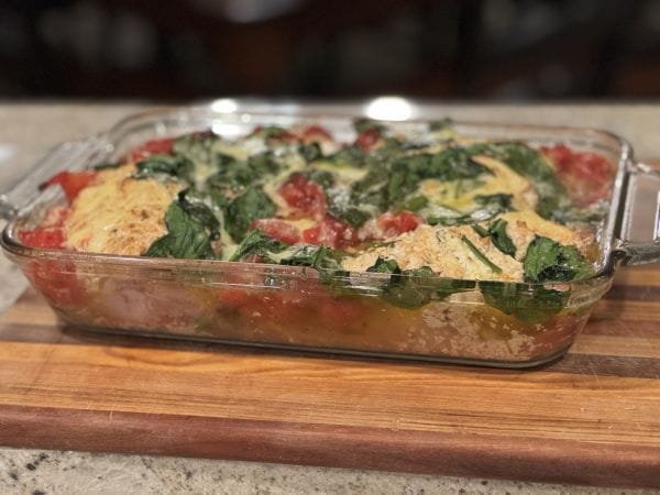 A baked casserole dish featuring chicken leg quarters, fresh spinach, diced tomatoes, and melted cheese on a wooden cutting board.