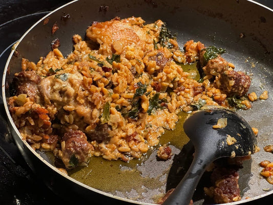 A skillet filled with Tuscan Chicken and Orzo, featuring golden-brown chicken thighs, Italian sausage, orzo pasta, and wilted greens, cooked in a creamy sauce.