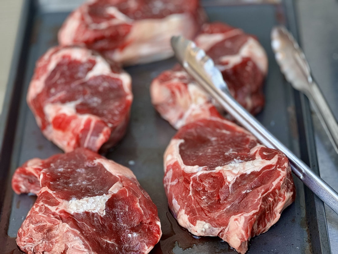 Close-up of raw grass-fed beef steaks on a grill pan with tongs, ready for cooking.