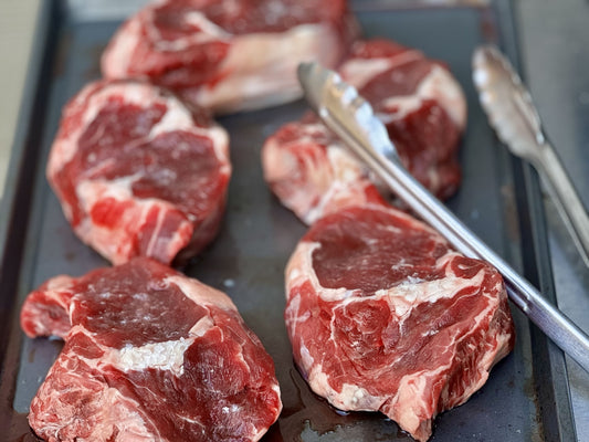 Close-up of raw grass-fed beef steaks on a grill pan with tongs, ready for cooking.