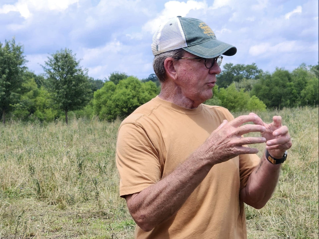 A man in a tan shirt and green cap discusses sustainable farming practices while standing in a grassy field at Tyner Pond Farm