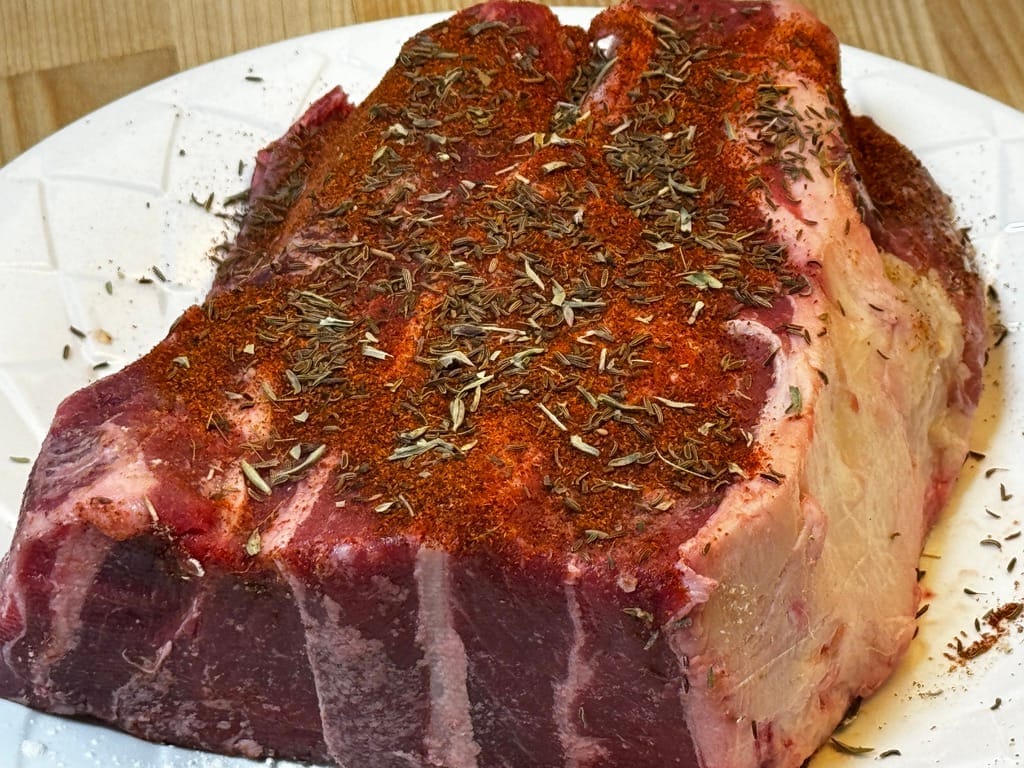 Raw grass-fed beef roast seasoned with herbs and spices on a white plate, highlighting its marbled texture and natural preparation