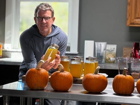 A man in glasses and a hoodie holds a mason jar filled with a golden liquid, standing behind a stainless steel counter with pumpkins and more jars of the same liquid. The setting is a kitchen with natural light coming through the window.