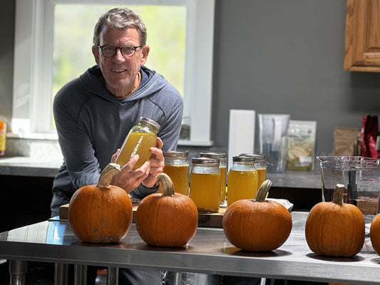A man holding a jar of homemade bone broth made from pasture-raised chickens, standing in a kitchen with pumpkins and jars of broth on the counter.