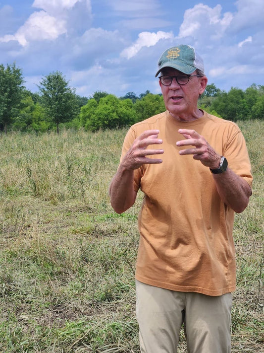 Chris Baggott of Tyner Pond Farm, wearing a tan shirt and green cap, gestures while explaining regenerative farming practices in a pasture with trees and a partly cloudy sky in the background.