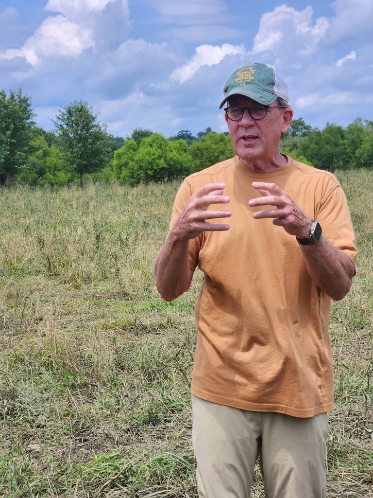 Chris Baggott of Tyner Pond Farm, wearing a tan shirt and green cap, gestures while explaining regenerative farming practices in a pasture with trees and a partly cloudy sky in the background.