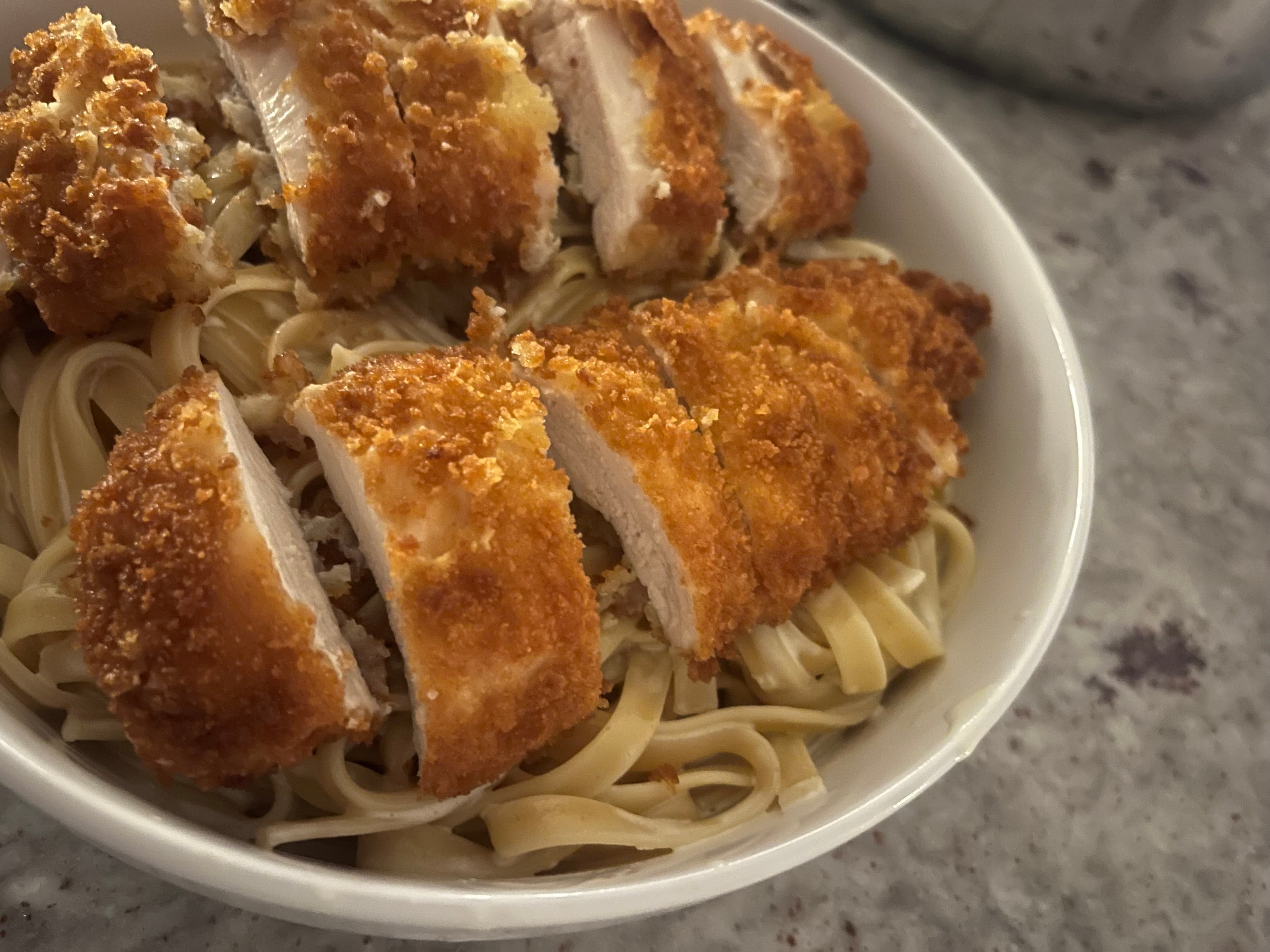 Lightly breaded Tyner Pond chicken breast sliced over pasta, topped with a simple pan sauce and chopped parsley in a shallow bowl.