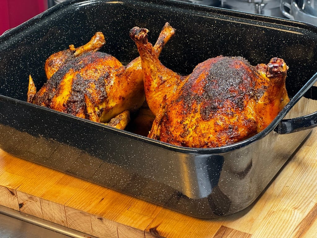 Two golden-brown pasture-raised chickens roasted with turmeric in a black roasting pan, showcasing the crispy skin and deep flavors of turmeric.