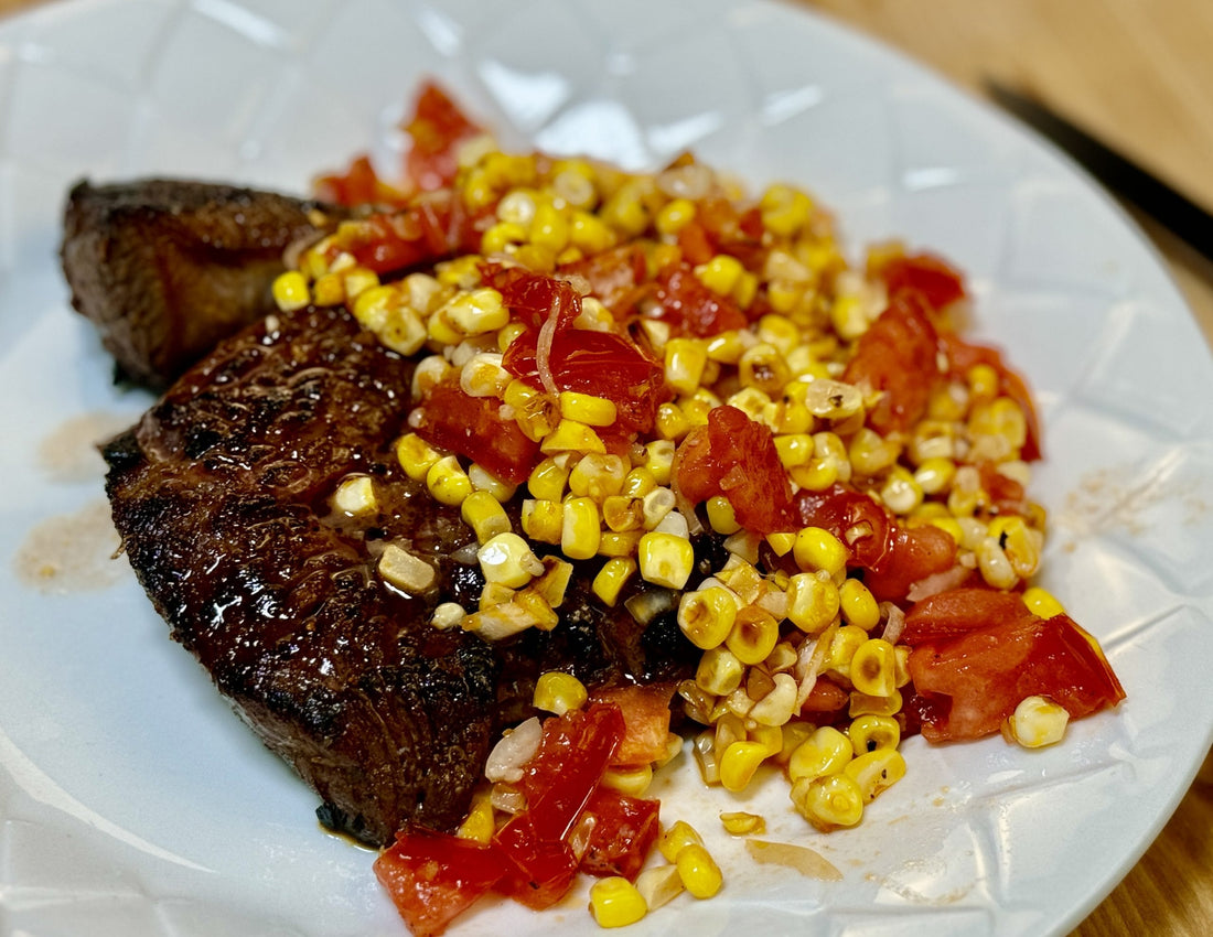 A plate of grass-fed sirloin topped with a fresh corn and heirloom tomato salad, showcasing a simple and delicious summer dish.