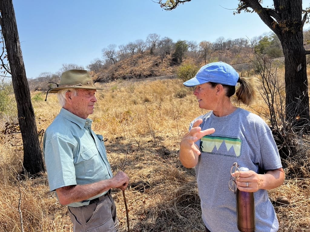 Allan Savory and Amy from Tyner Pond Farm stand in a dry grassy field in Zimbabwe, discussing sustainable grazing and land management.