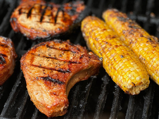 Two thick-cut pork chops, marked with grill lines and glistening with marinade, cook beside ears of corn, their kernels beginning to char. The scene captures the essence of a summer evening meal, simple yet satisfying.