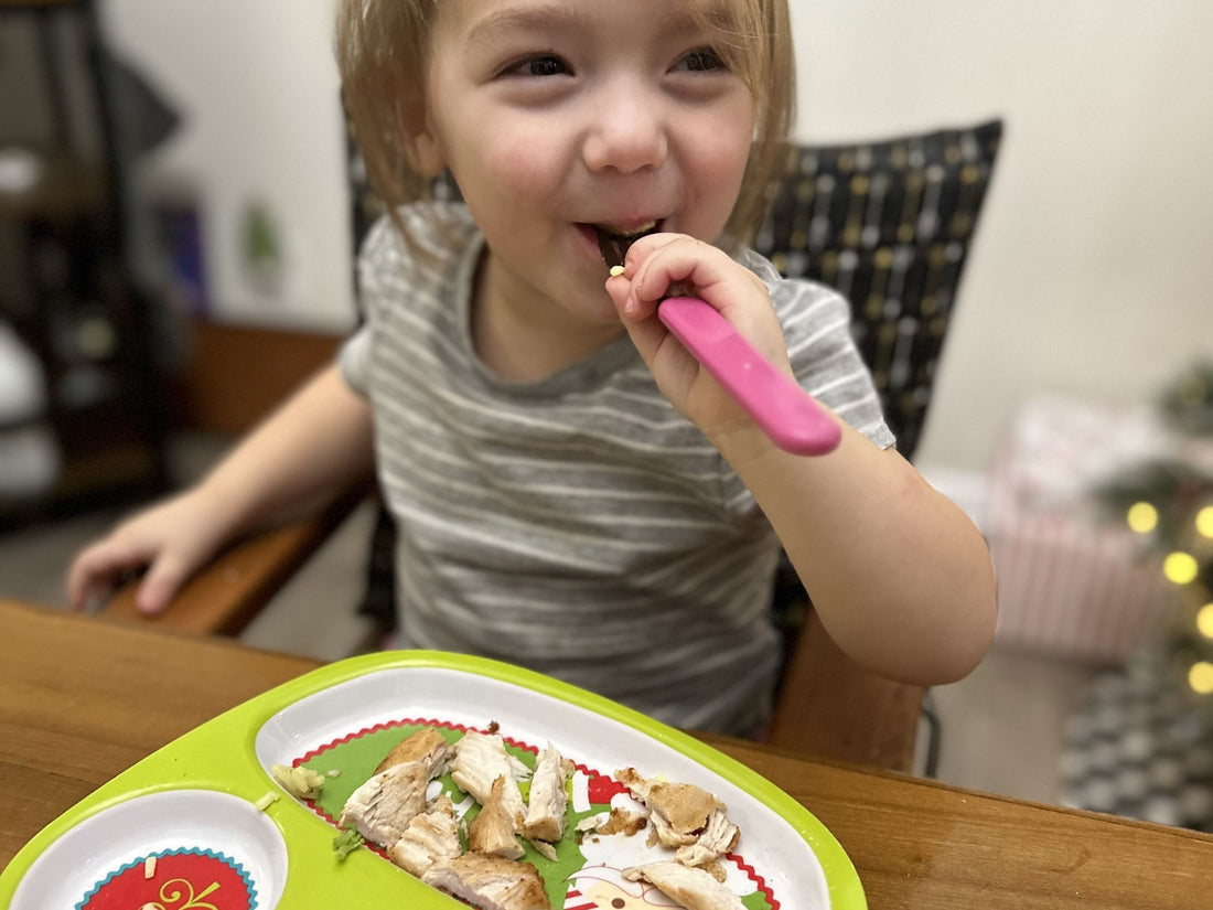 A toddler sitting at a wooden table, smiling while using a pink fork to eat pieces of cooked Tyner Pond Farm pasture-raised whole chicken. The food is arranged on a green and red sectioned plate, with a blurred background featuring Christmas decorations.