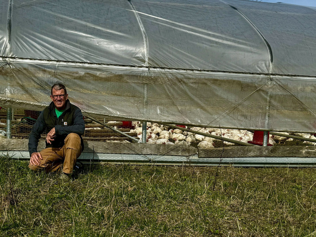 A farmer kneeling in front of a mobile pasture shelter with pasture-raised chickens visible inside, set on a green, grassy field