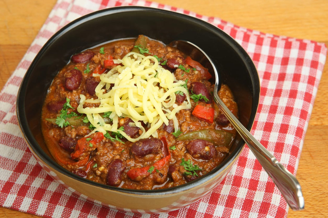 A bowl of hearty chili topped with shredded cheese and fresh parsley, served in a black bowl with a spoon, placed on a red-and-white checkered napkin.
