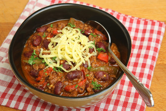 A bowl of hearty chili topped with shredded cheese and fresh parsley, served in a black bowl with a spoon, placed on a red-and-white checkered napkin.