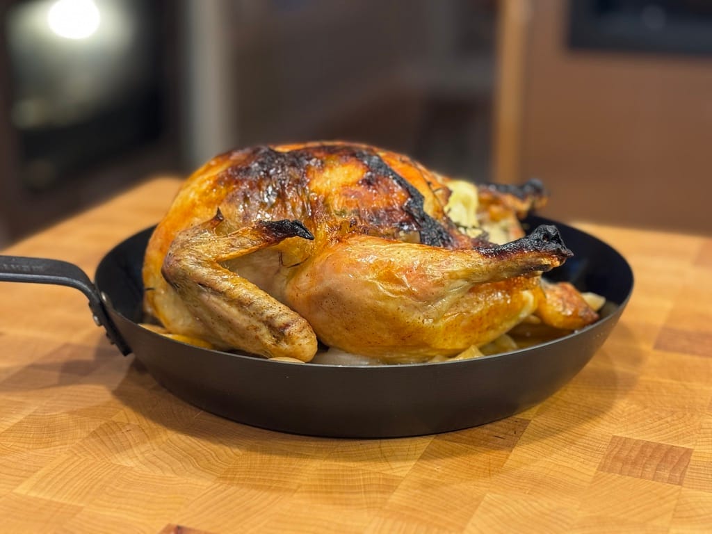 A perfectly roasted pasture-raised chicken cooked in a Dutch oven, golden brown and resting on a wooden cutting board, surrounded by its natural juices.