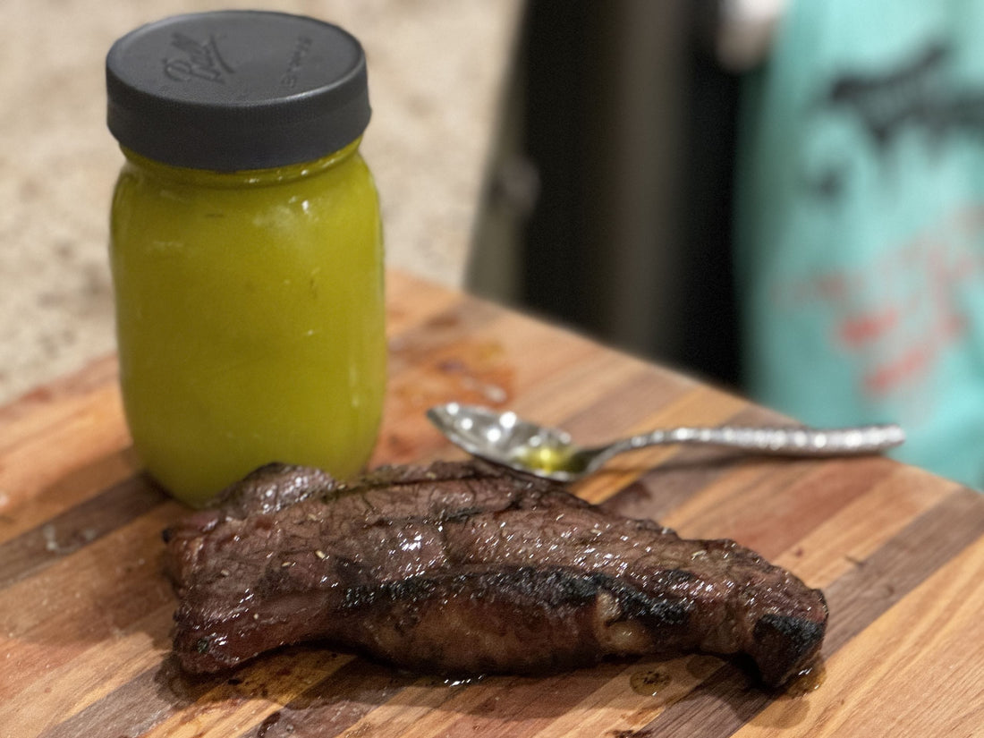 A jar of herb-infused steak oil next to a freshly cooked grass-fed steak on a wooden cutting board.