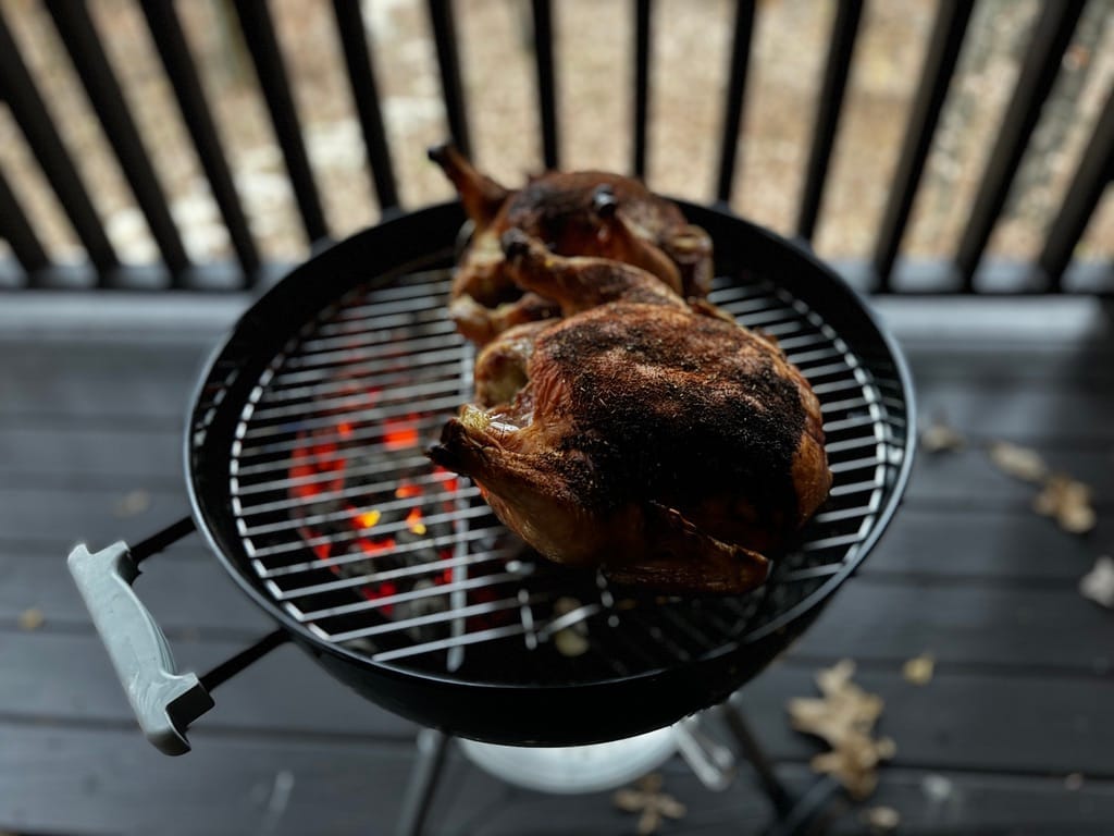 Two whole pasture-raised chickens grilling on a charcoal grill, with a rich brown finish on each
