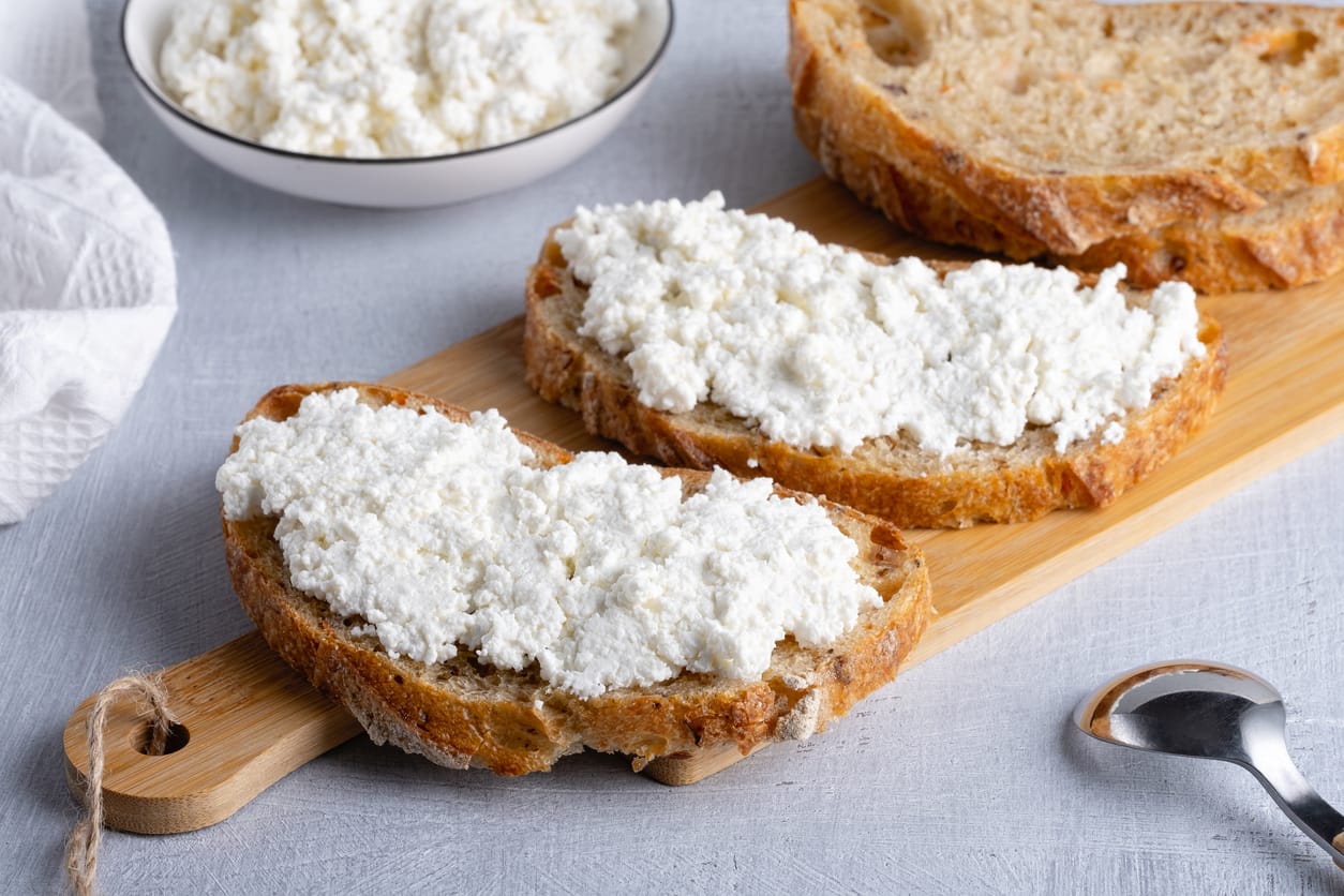 Two slices of crusty bread topped with fresh homemade cottage cheese on a wooden board, with a bowl of cottage cheese in the background