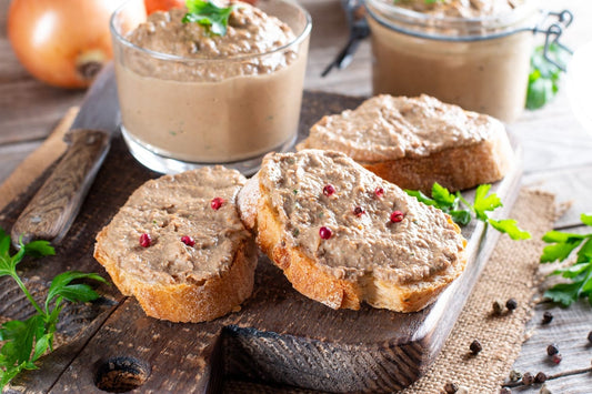 Pasture-raised chicken liver pâté served on rustic bread with pepper garnish, surrounded by fresh herbs