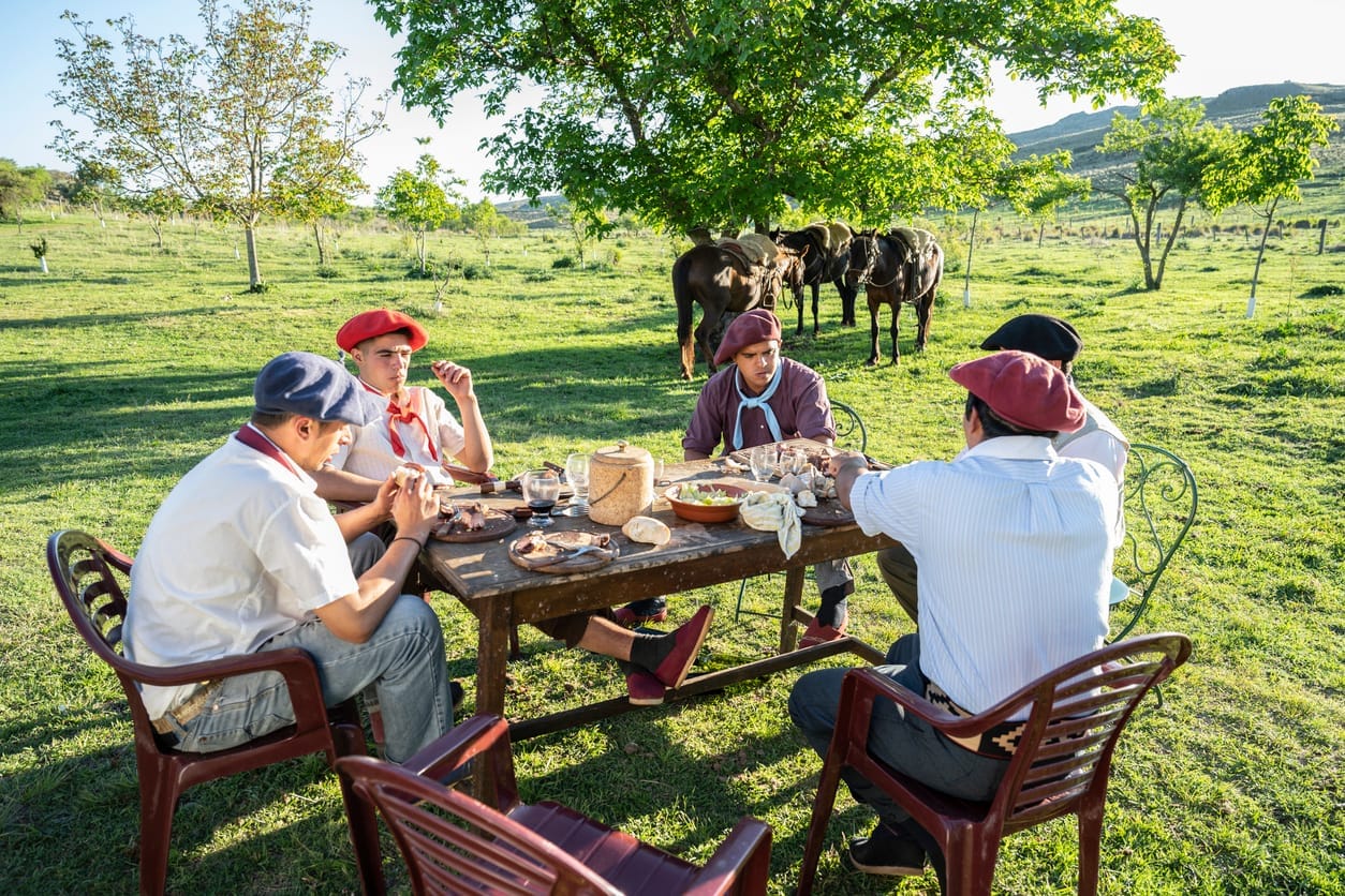 Gauchos gathered around an outdoor table in the Argentine countryside, sharing a rustic meal with horses in the background.