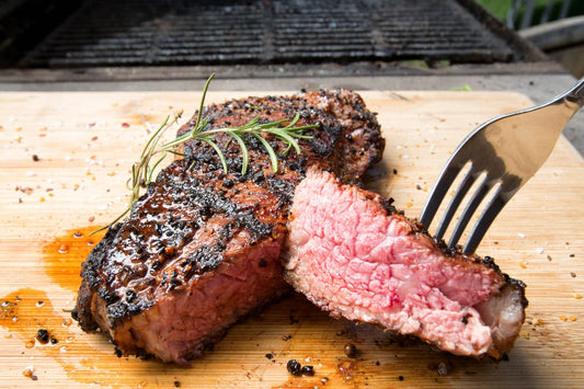 A freshly grilled, grass-finished New York strip steak, sliced to reveal a juicy medium-rare interior, served on a wooden cutting board with coarse black pepper, salt, and a sprig of rosemary.