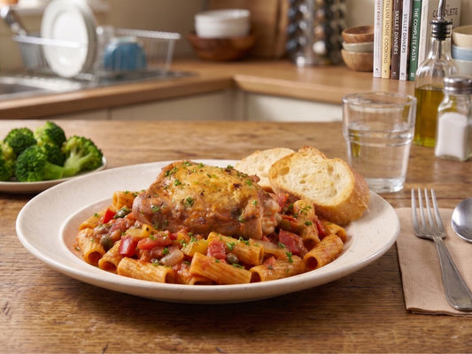 A perfectly seared, crispy pasture-raised chicken thigh served over rigatoni pasta in a rich tomato Puttanesca sauce, accompanied by sides of steamed broccoli and crusty French bread on our counter