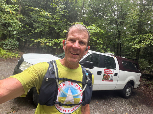 Chris Baggott smiling in a lime-green shirt and hydration pack beside a Tyner Pond Farm pickup in the woods.