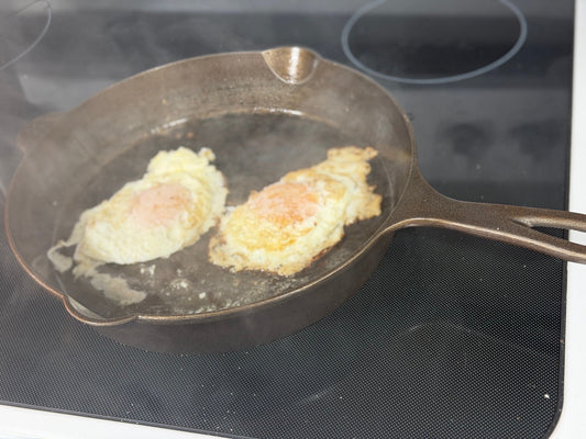 Two fried eggs in a seasoned cast-iron skillet on a black glass stovetop, with the handle extending to the right.