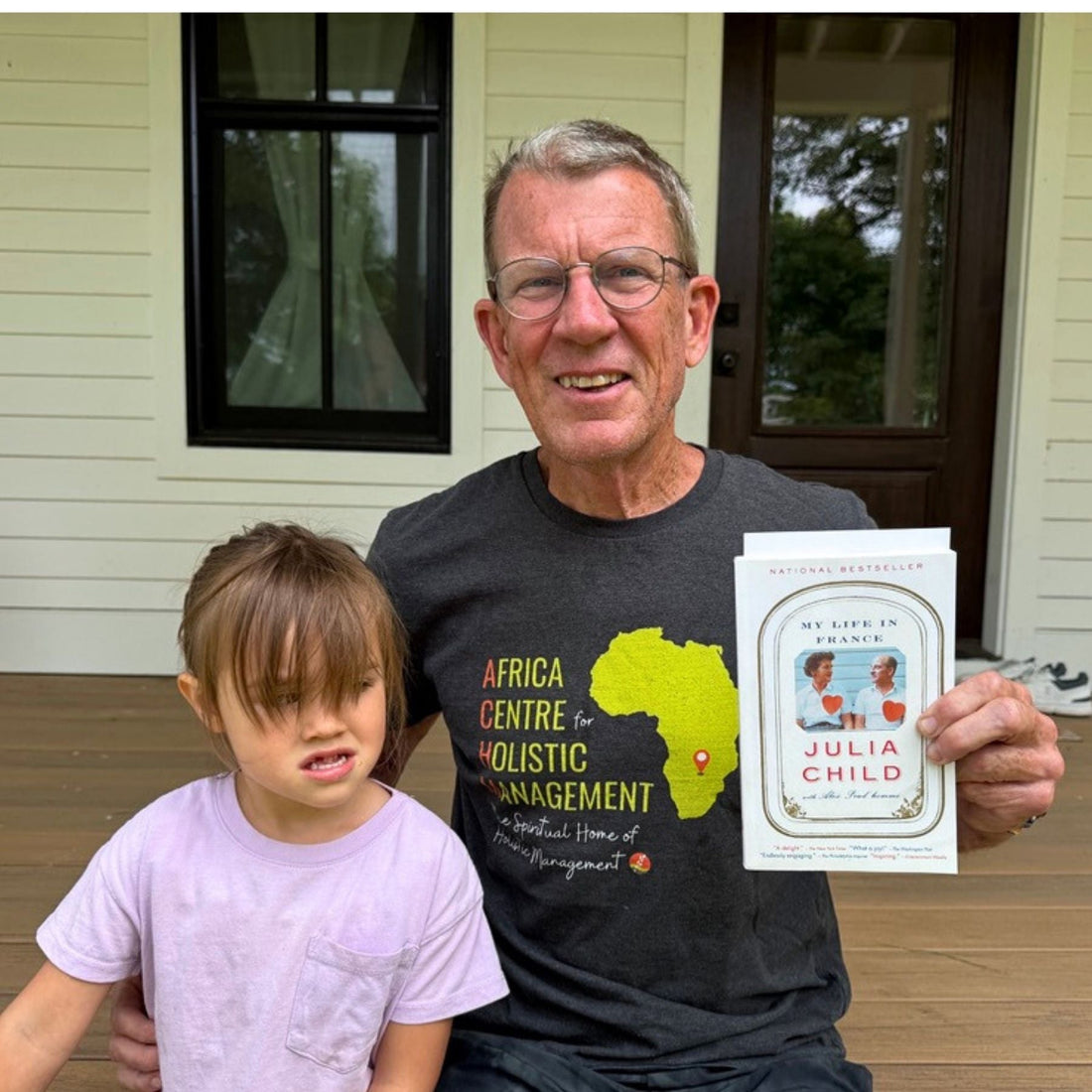 Chris Baggott holding Julia Child’s My Life in France on a farmhouse porch with a child beside him.