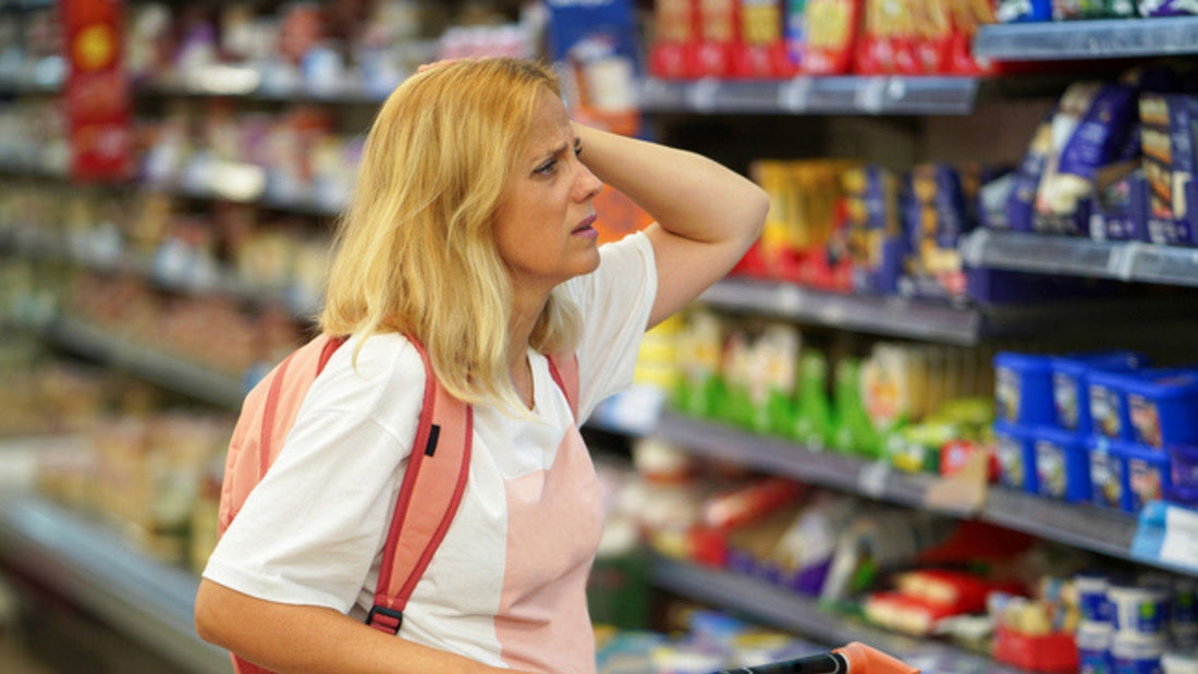 Mother standing in a grocery store aisle with a shopping cart, appearing overwhelmed while deciding what to buy