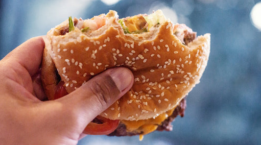 A hand holding a half-eaten fast-food cheeseburger on a sesame seed bun, a meal built from layered salt, fat, and refined carbohydrates.