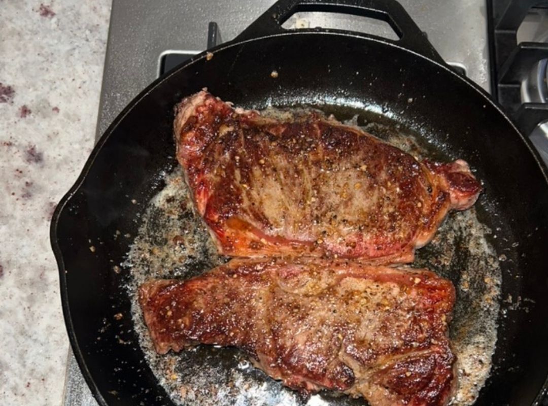 Grass-fed New York Strip steaks searing in a vintage cast iron skillet.