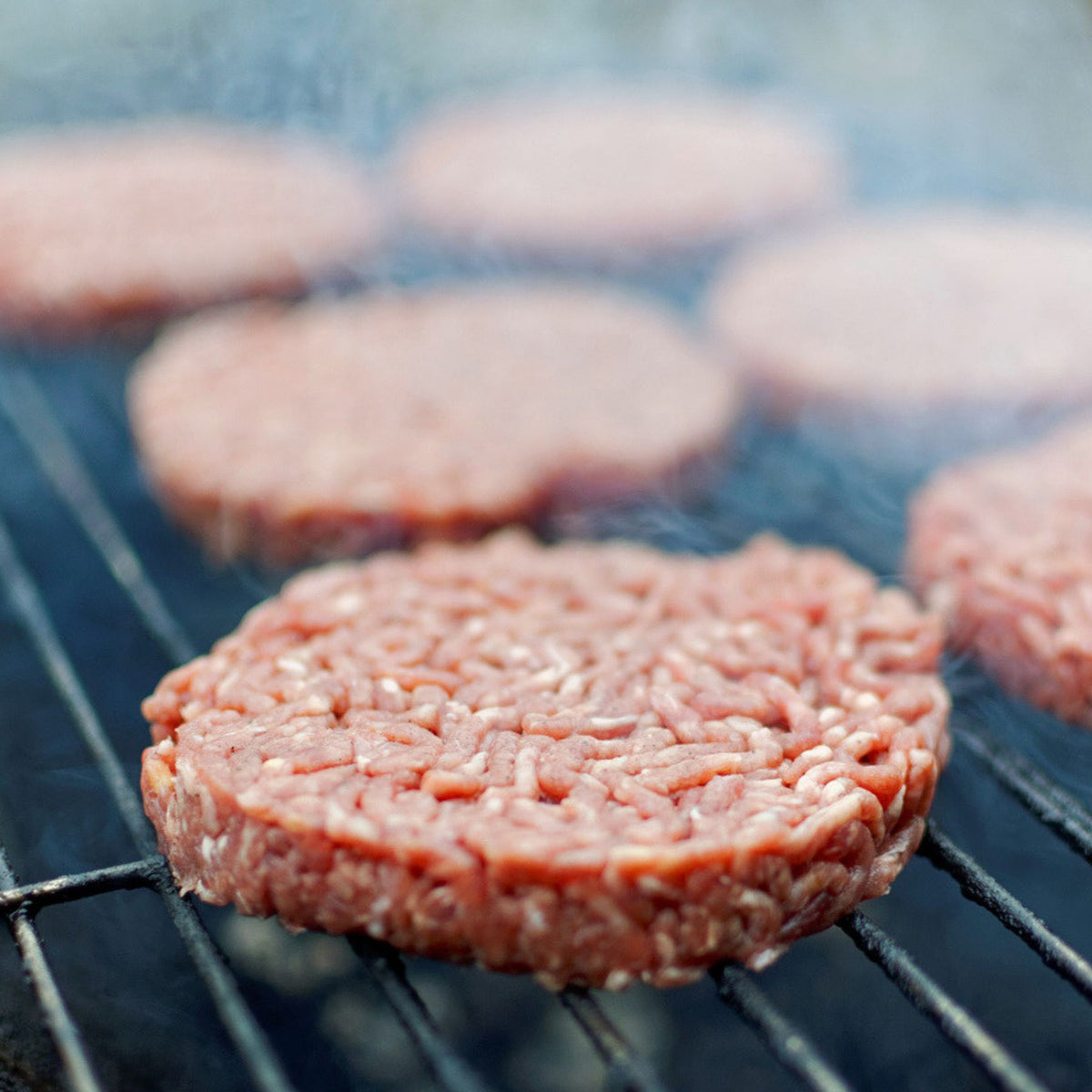 100% grass-fed beef patties cooking on a grill with smoke rising around the burgers.