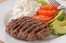 A close-up of a grilled beef patty on a white plate, served with a scoop of white cheese, sliced tomatoes, avocado, and a fork.
