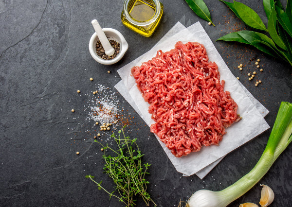 Ground grassfed beef on a piece of paper with herbs and spices on a dark surface