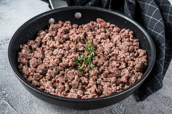 Grassfed Ground beef in a black frying pan on a gray surface with a checkered towel.