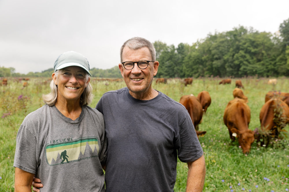 Chris and Amy Baggott of Tyner Pond Farm standing in a field with cows in the background