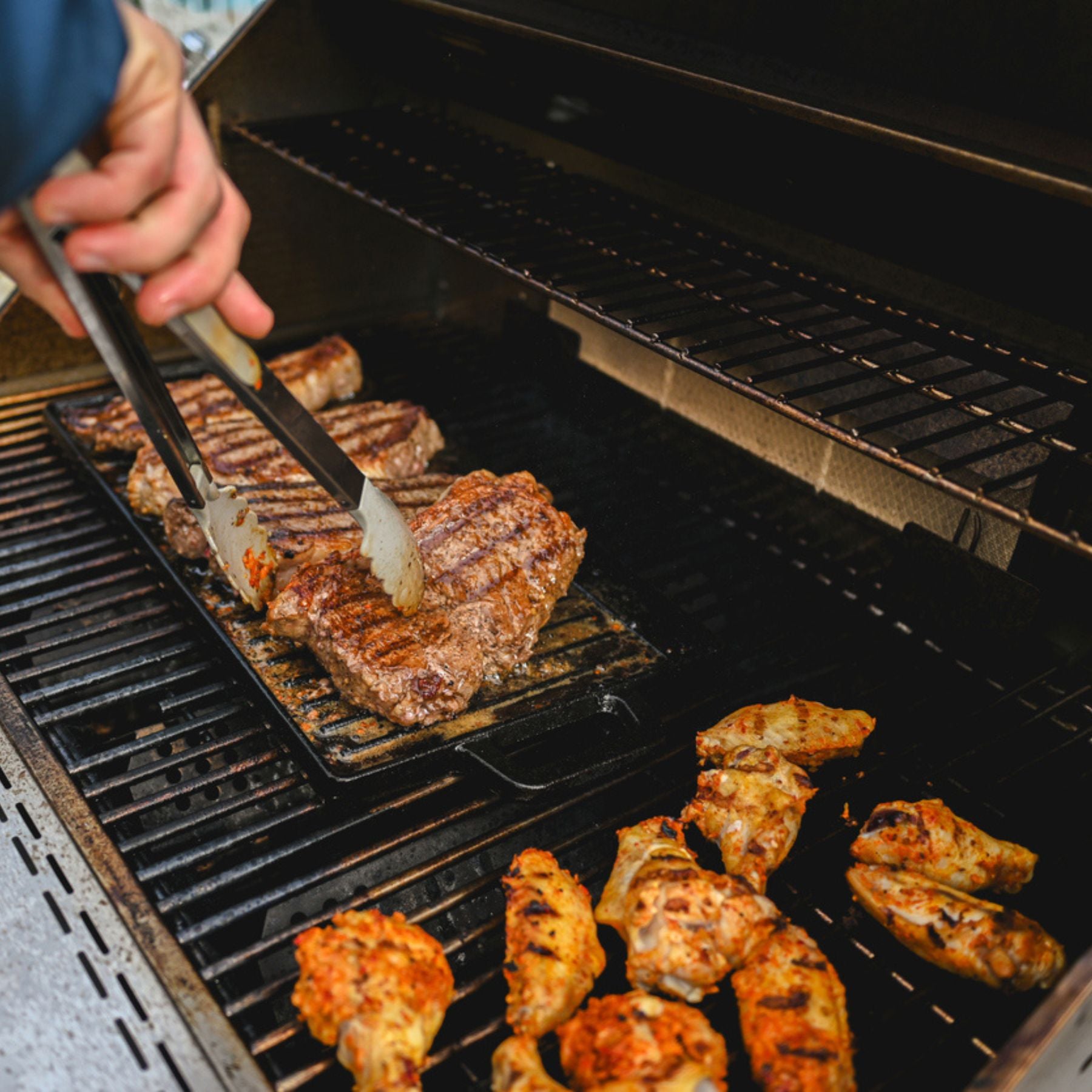 A close-up of a hand using metal tongs to flip a grilled grassfed steak on a black grill grate, with chicken pieces cooking nearby and a visible hand in frame.