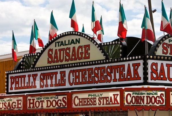 Food stand sign advertising Italian sausage, Philly style cheesesteak, and other food items with Italian flags in the background.