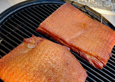 Two pork belly slabs smoking on Weber grill, being prepared for homemade bacon
