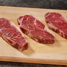 Three raw grassfed beef steaks with visible marbling rest on a light wood cutting board against a dark countertop.